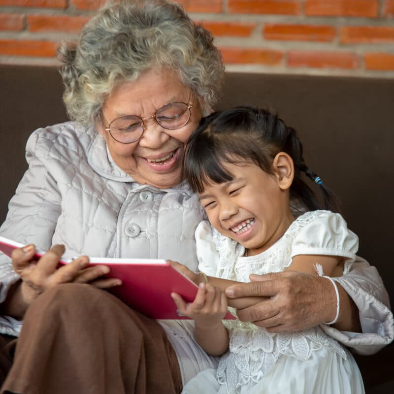 An elderly woman smiling and laughing with a little girl while they read a children's book.